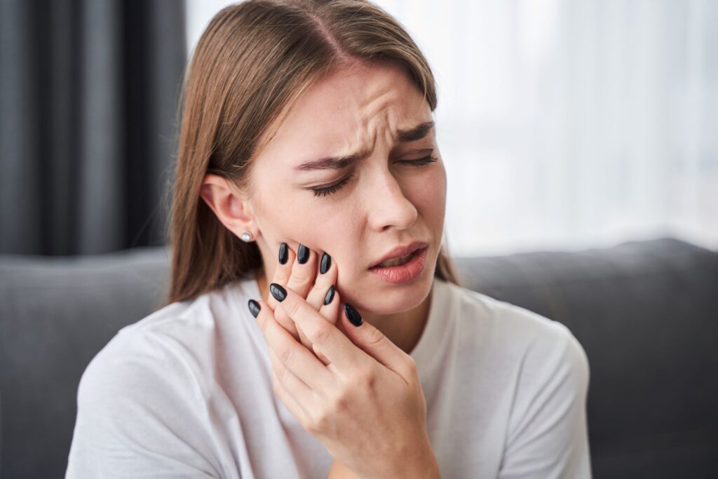 Woman with brown hair and dark nails pressing hands to sore jaw